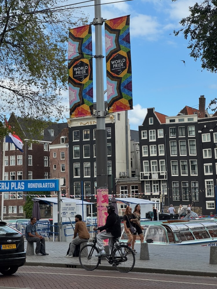 A cyclists bikes passed a lamp post with the World Pride banner for Amsterdam 2026.