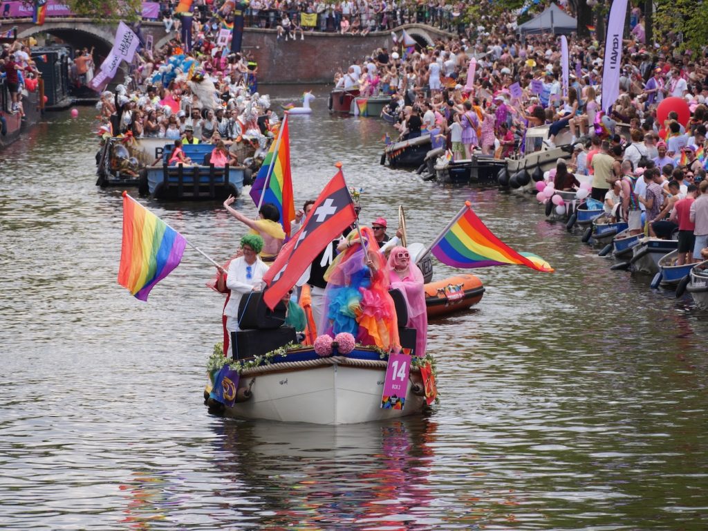 A small boat with multiple rainbow flags and one Amsterdam XXX flag during the Amsterdam World Pride 2026 Canal Parade.