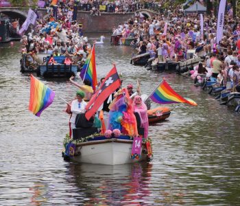 A small boat with multiple rainbow flags and one Amsterdam XXX flag during the Amsterdam World Pride 2026 Canal Parade.