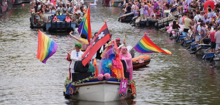 A small boat with multiple rainbow flags and one Amsterdam XXX flag during the Amsterdam World Pride 2026 Canal Parade.